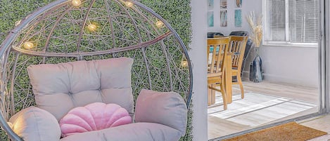 A room with a hanging chair, a green wall with a neon sign, and a dining area with chairs and a table.