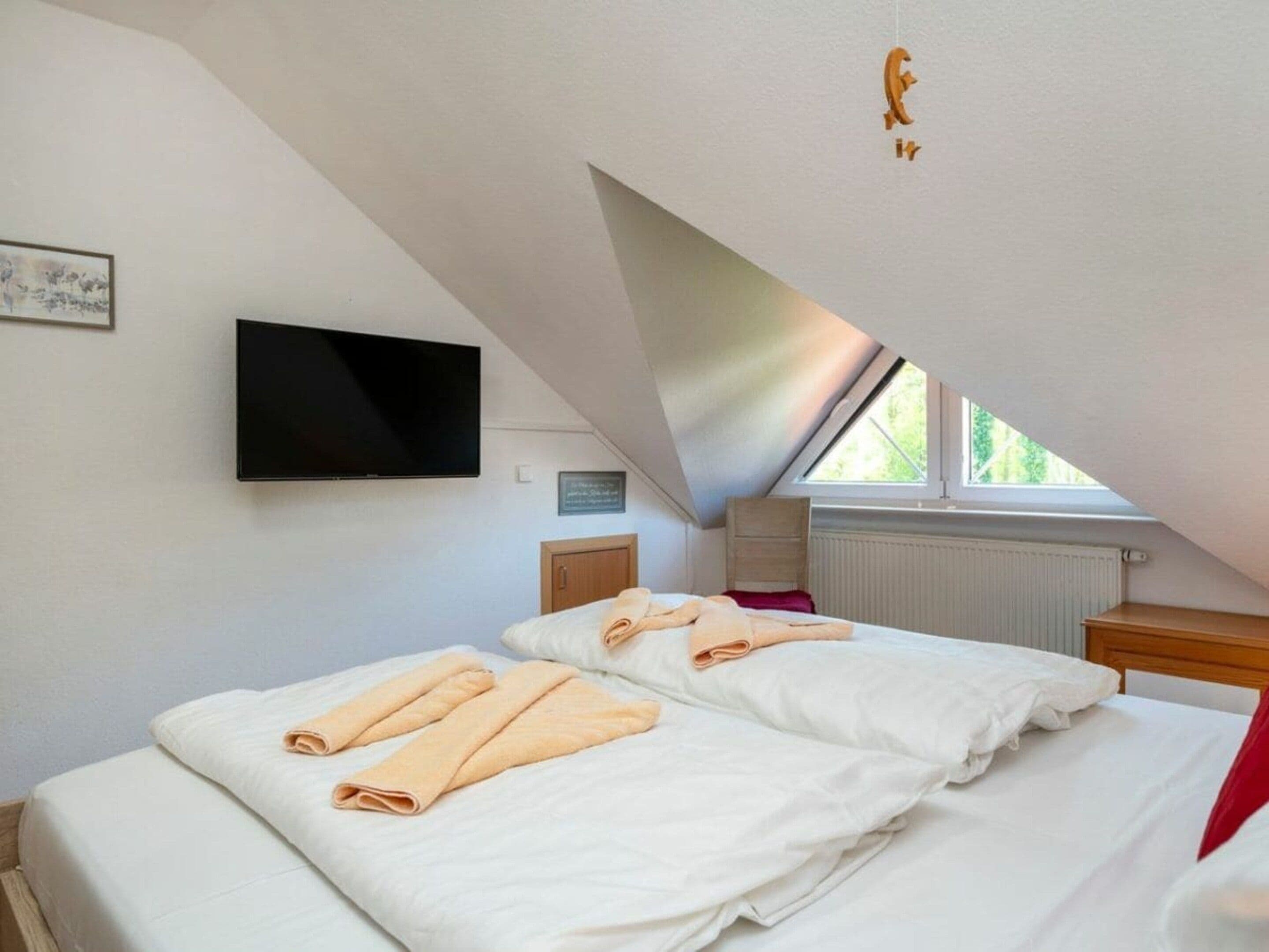A bedroom with a sloped ceiling, a flat-screen TV mounted on the wall, a bed with white linens and rolled towels, a window with a view of greenery, and a radiator below the window.