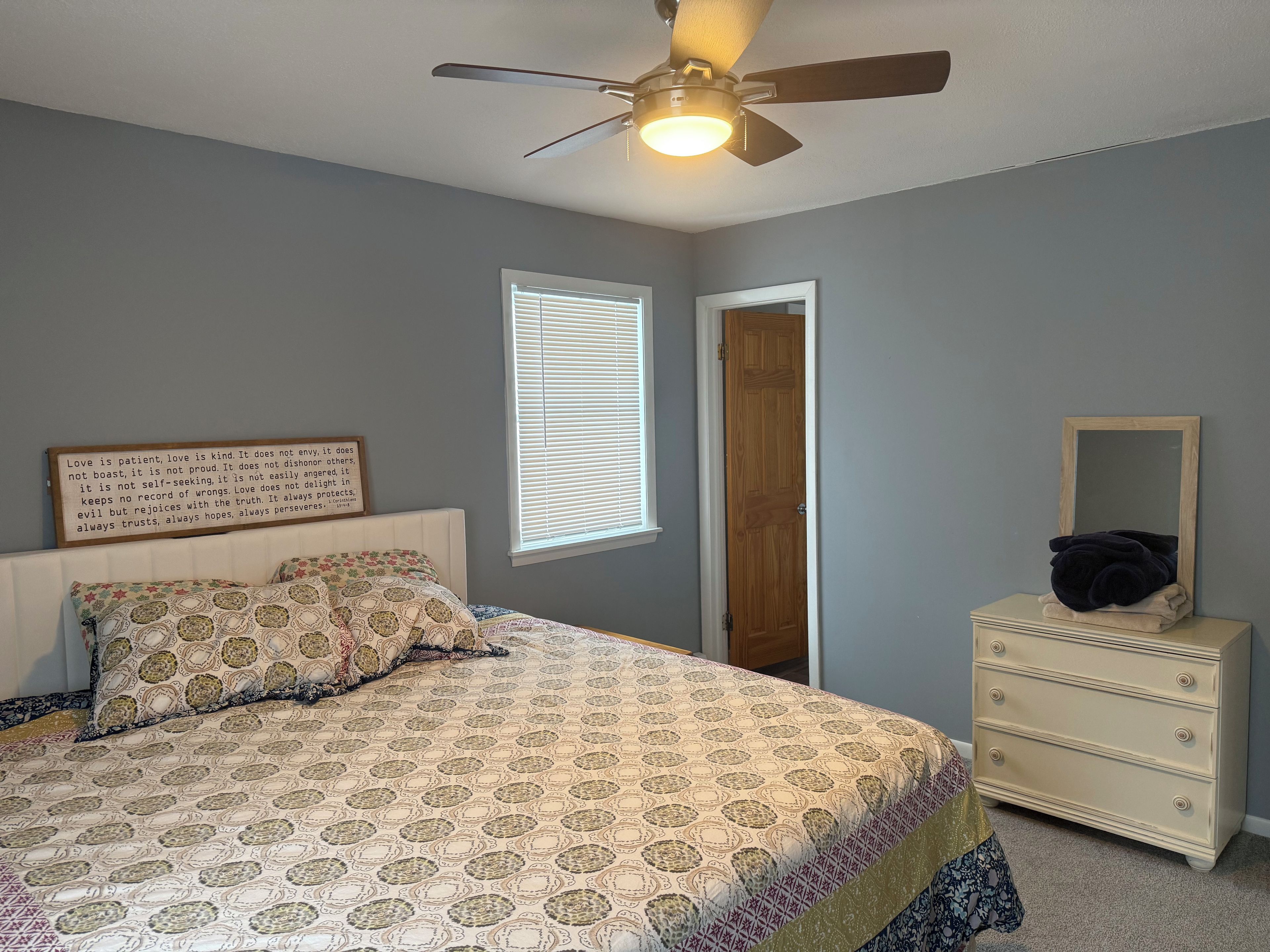 A bedroom with a bed, a dresser, a ceiling fan, a window with blinds, and a motivational quote on the wall.