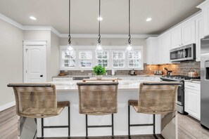 A modern kitchen with a central island, three wicker bar stools, pendant lighting, and stainless steel appliances. - Come & Sea by Stay on 30a (Inlet Beach)