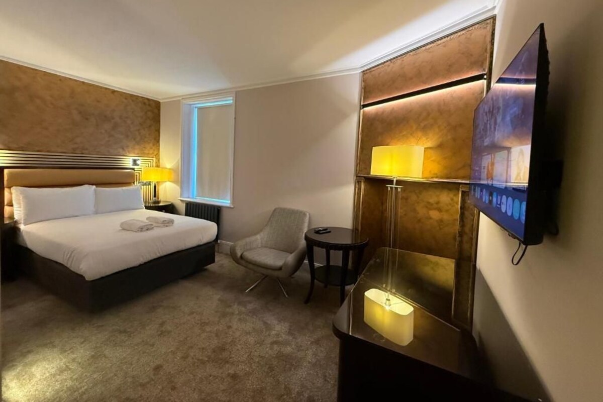 A hotel room with a bed, a chair, a small table, a TV, and a wall-mounted lamp.