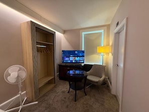 A hotel room with a TV, a chair, a desk, a lamp, and a closet.