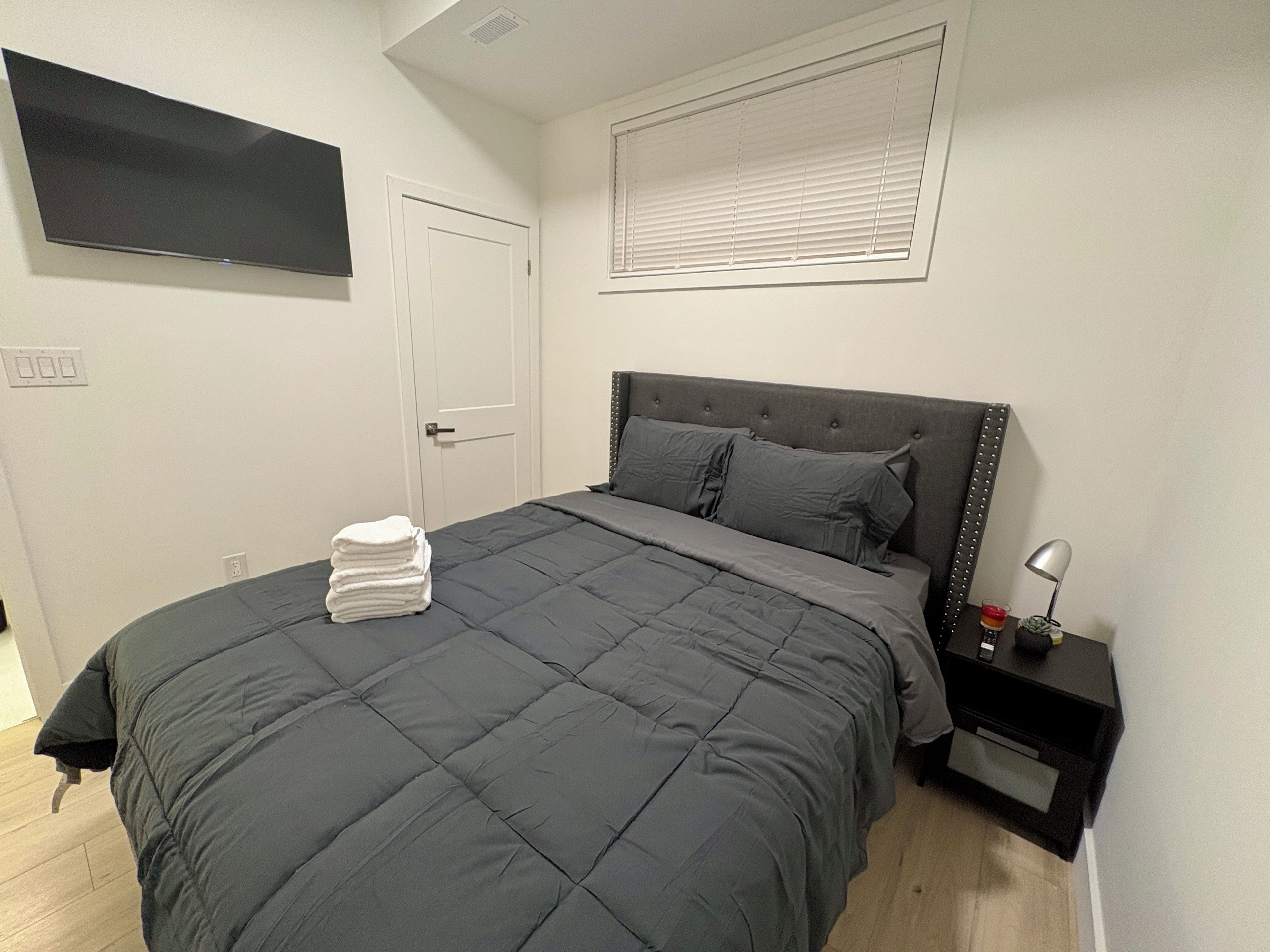 A bedroom with a bed, a bedside table with a lamp, a wall-mounted TV, and a door.