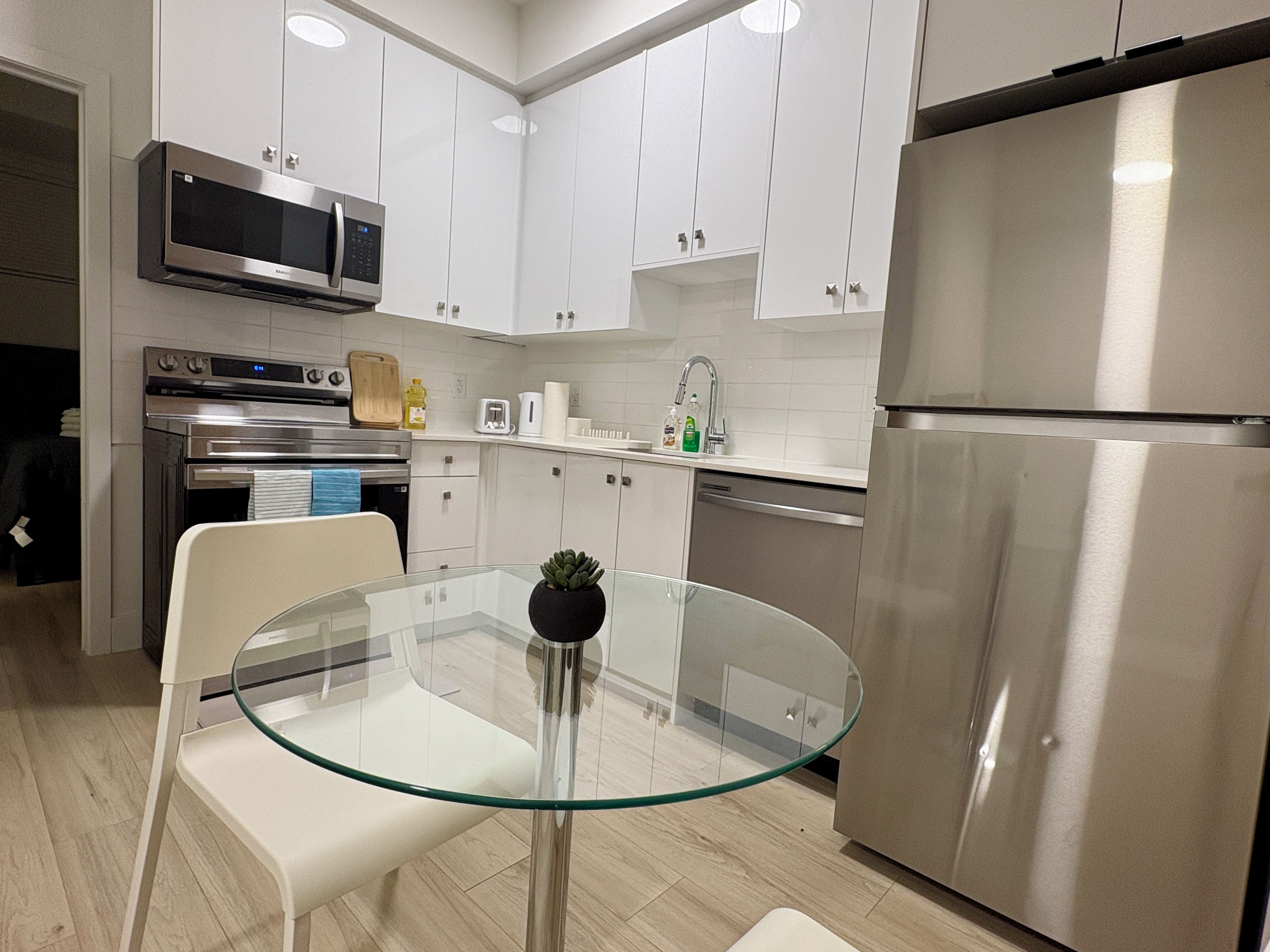 A modern kitchen with white cabinets, stainless steel appliances, and a glass table with a potted plant.