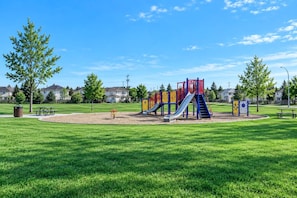 Ein Spielplatz mit Rutschen und Klettergerüsten, umgeben von einer Wiese und Bäumen, mit Häusern im Hintergrund.