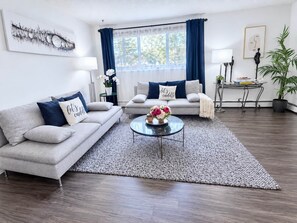 Ein Wohnzimmer mit einem grauen Sofa, einem Couchtisch mit Blumen, einem blauen Vorhang und einem Gemälde an der Wand.