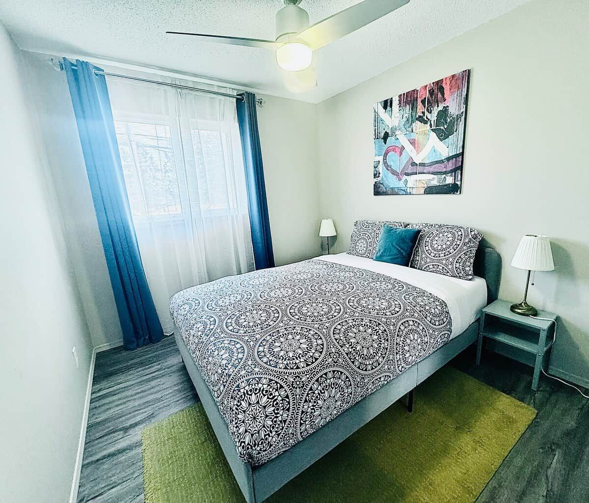 Une chambre d’hôtel avec un lit, une table de chevet, une lampe, un tableau au mur et une fenêtre avec des rideaux.
