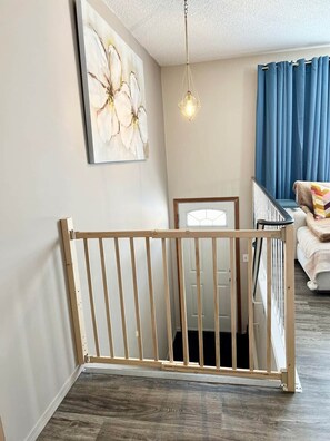 A room with a wooden crib, a wall-mounted artwork, a hanging light fixture, and a view of a bed with blue curtains. - Family Baby&Kid Friendly*Close toUofA*Kingbed*3BR* (Edmonton)