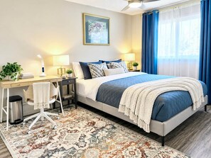 A modern hotel room with a large bed, a desk with a chair, a small table with a lamp, a potted plant, and a patterned rug. - Family Baby&Kid Friendly*Close toUofA*Kingbed*3BR* (Edmonton)