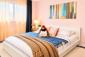 A neatly made bed with a plush toy, a bedside table with a lamp, and a colorful abstract painting on the wall. - Funky family-friendly 3BR, king bed near U of A (Edmonton)