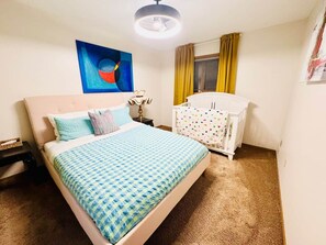 Ein Schlafzimmer mit einem Bett, einem Babybett, einem Nachttisch, einer Lampe und einem Bild an der Wand.
