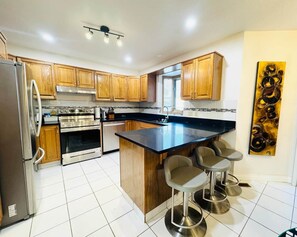 A modern kitchen with wooden cabinets, stainless steel appliances, a black countertop island with bar stools, and a decorative wall piece. - KidsParadise*Kingbed*NearWEM*BabyFamilyKid Frienly (Edmonton)