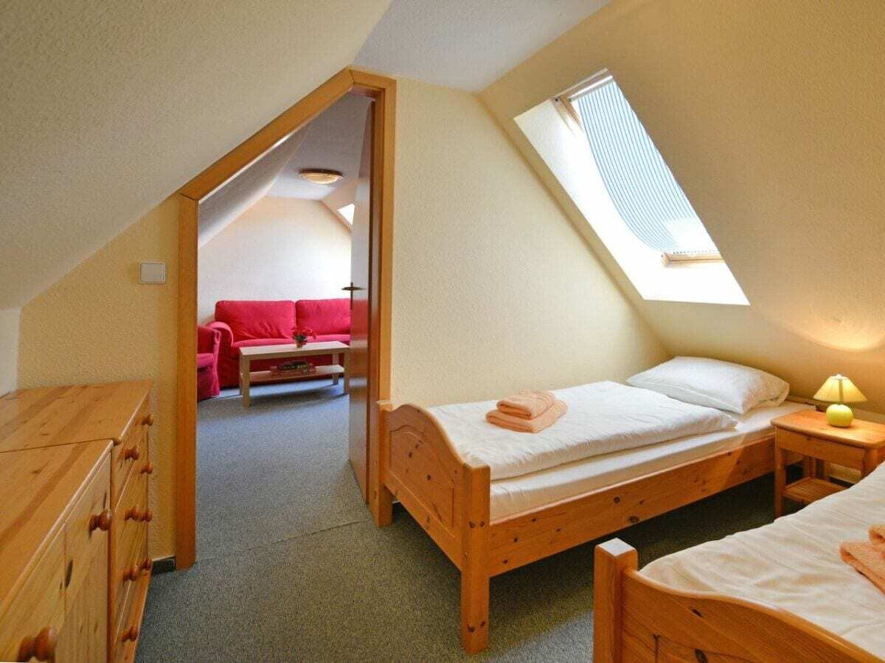 A bedroom with a sloped ceiling, a wooden bed, a nightstand, a lamp, and a view of a red sofa in the adjacent room.