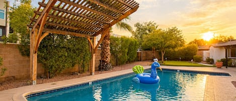A swimming pool with a wooden pergola, palm trees, and a peacock float.