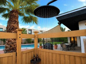 A wooden deck with a built-in showerhead, a palm tree, and a pool in the background. - Wellness Retreat: Pool, Hot Tub, Mini Golf, Outdoor Shower + Old Town Scottsdale (Scottsdale)