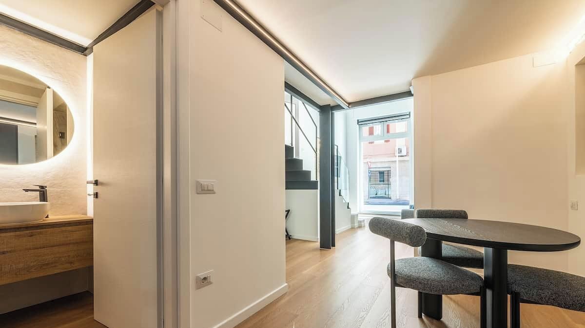 Interior moderno com um banco de madeira e uma mesa redonda, uma porta branca e vista para uma escadaria que leva a uma varanda.