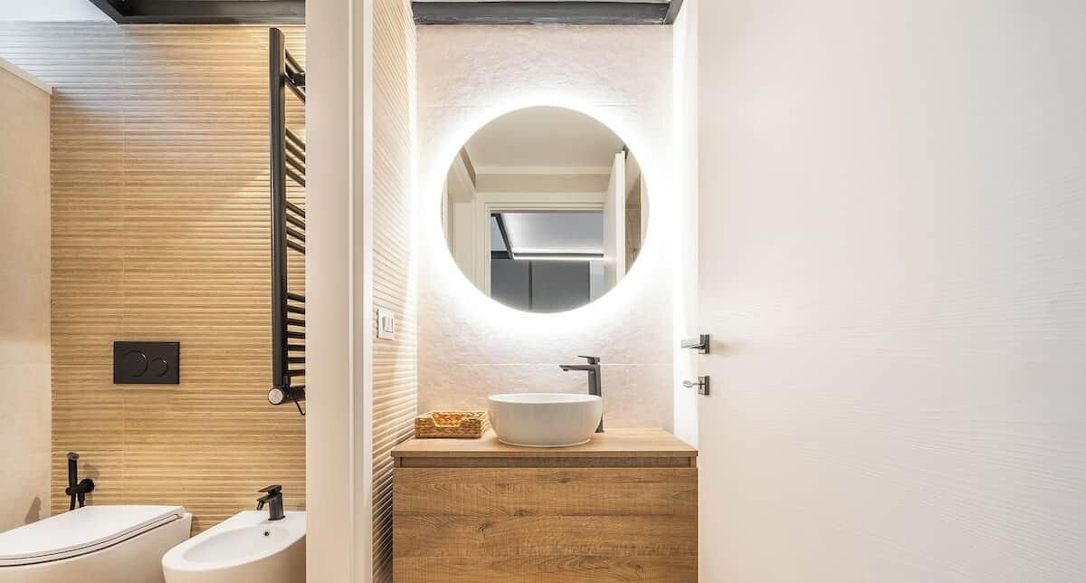 A modern bathroom with a wooden vanity, a round mirror with built-in lighting, and two sinks.