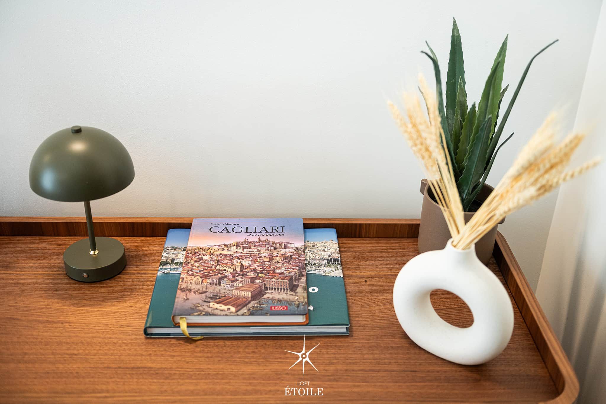 A book titled 'CAGLIARI' on a wooden tray, a desk lamp, and a white vase with dried plants.