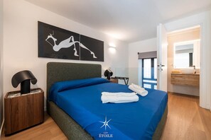 Quarto de hotel moderno com cama, mesa de cabeceira, luminária, obra de arte na parede e banheiro visível através de uma porta aberta.