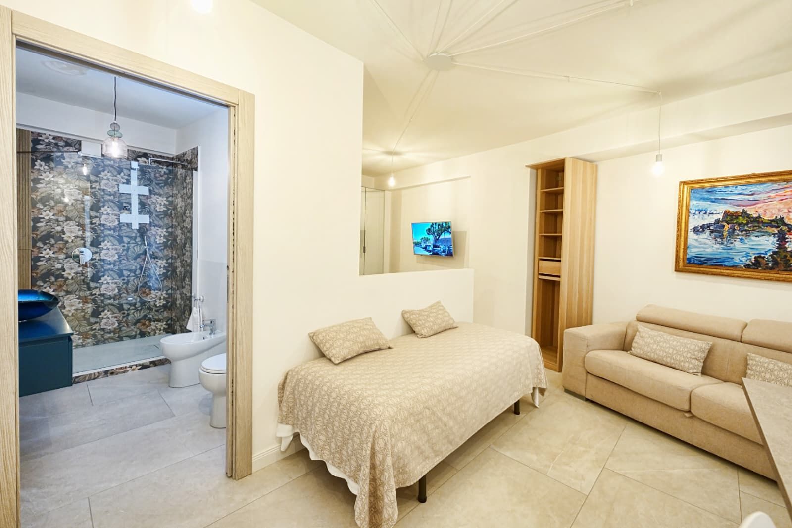 Un dormitorio con cama, sofá, televisión, un cuadro y un baño con ducha e inodoro.