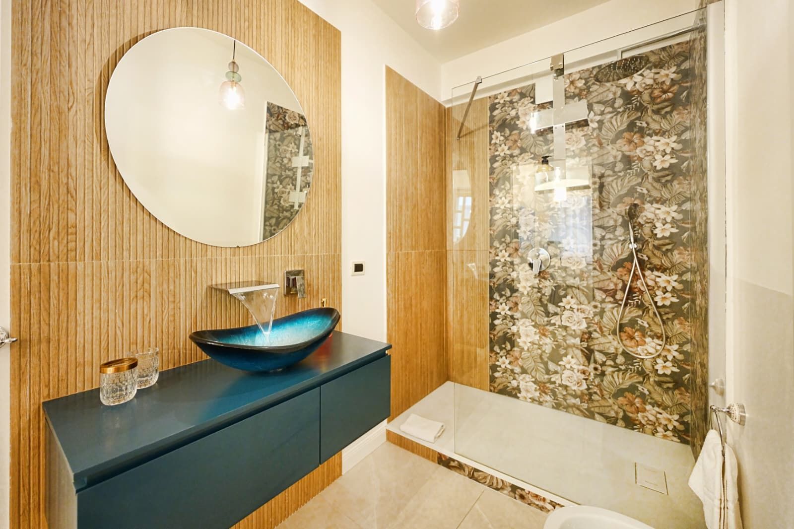 Un bagno moderno con uno specchio rotondo, un lavabo a forma di vaso blu, una doccia con carta da parati floreale e pannelli in legno.