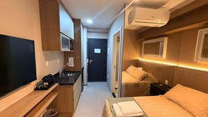 Habitación de hotel compacta con cama, televisión, kitchenette y equipo de aire acondicionado.