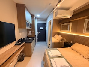 A compact hotel room with a bed, a TV, a kitchenette, and an air conditioning unit. - New Studio in Jardins | 2 Blocks from Paulista | Pool, Gym & Coworking (São Paulo)
