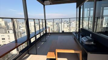 Un balcon situé dans un immeuble de grande hauteur, offrant une vue sur le paysage urbain, avec un banc en bois et une petite table sur laquelle se trouve une tasse.