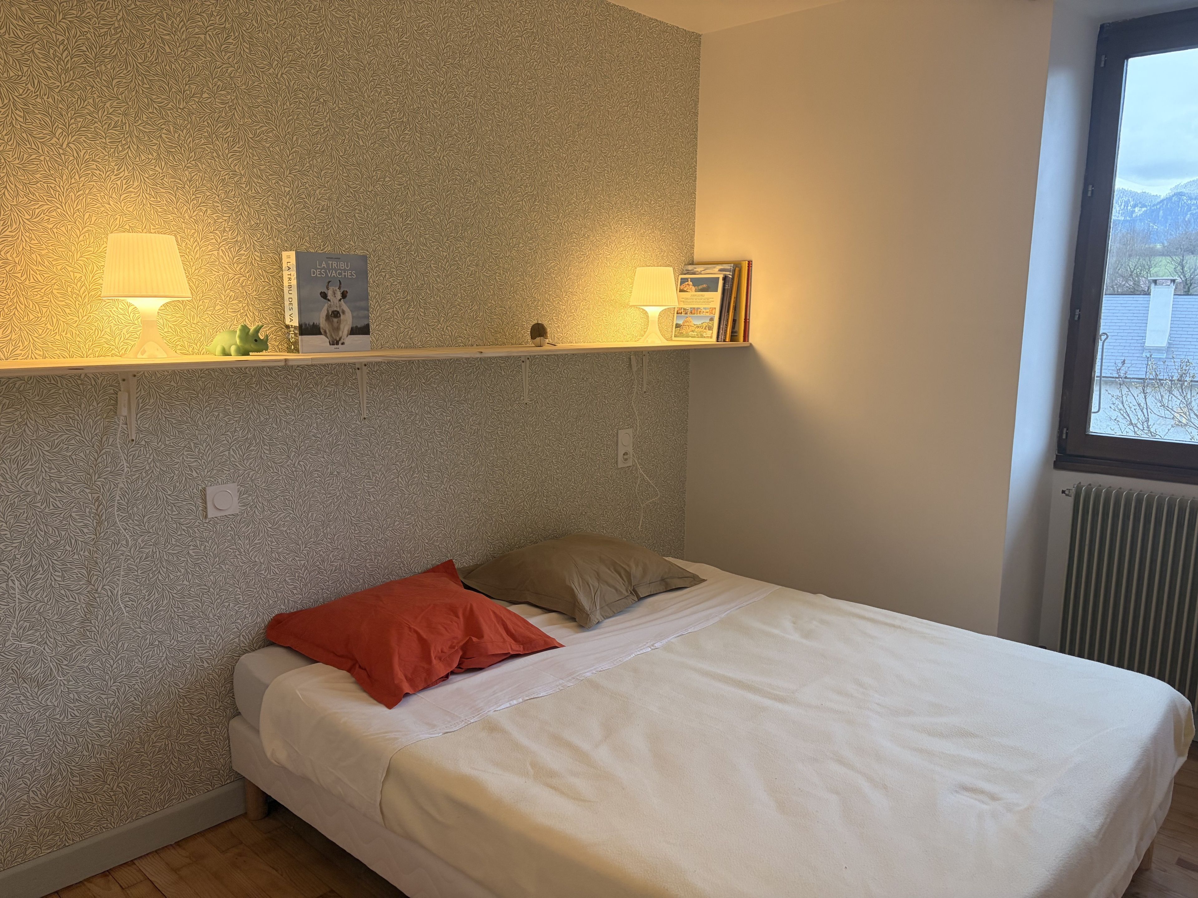 Een slaapkamer met een bed, kussens, een plank met decoratieve objecten, een raam en een radiator.