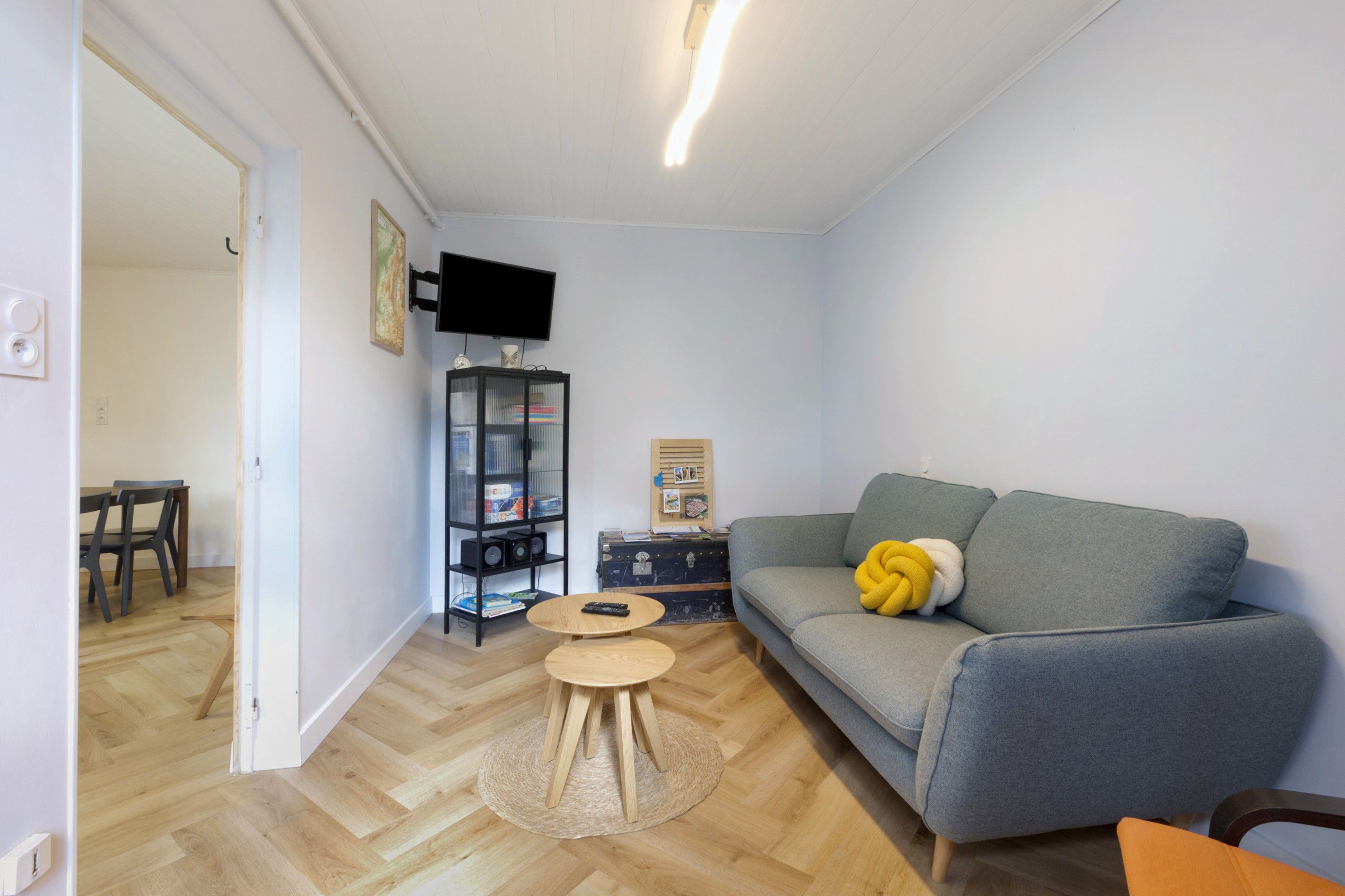 Een woonkamer met een grijze bank, een kleine ronde tafel, een boekenkast en een flatscreen televisie aan de muur.