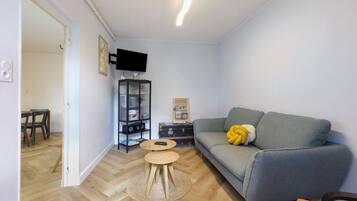 Een woonkamer met een grijze bank, een kleine ronde tafel, een boekenkast en een flatscreen televisie aan de muur.
