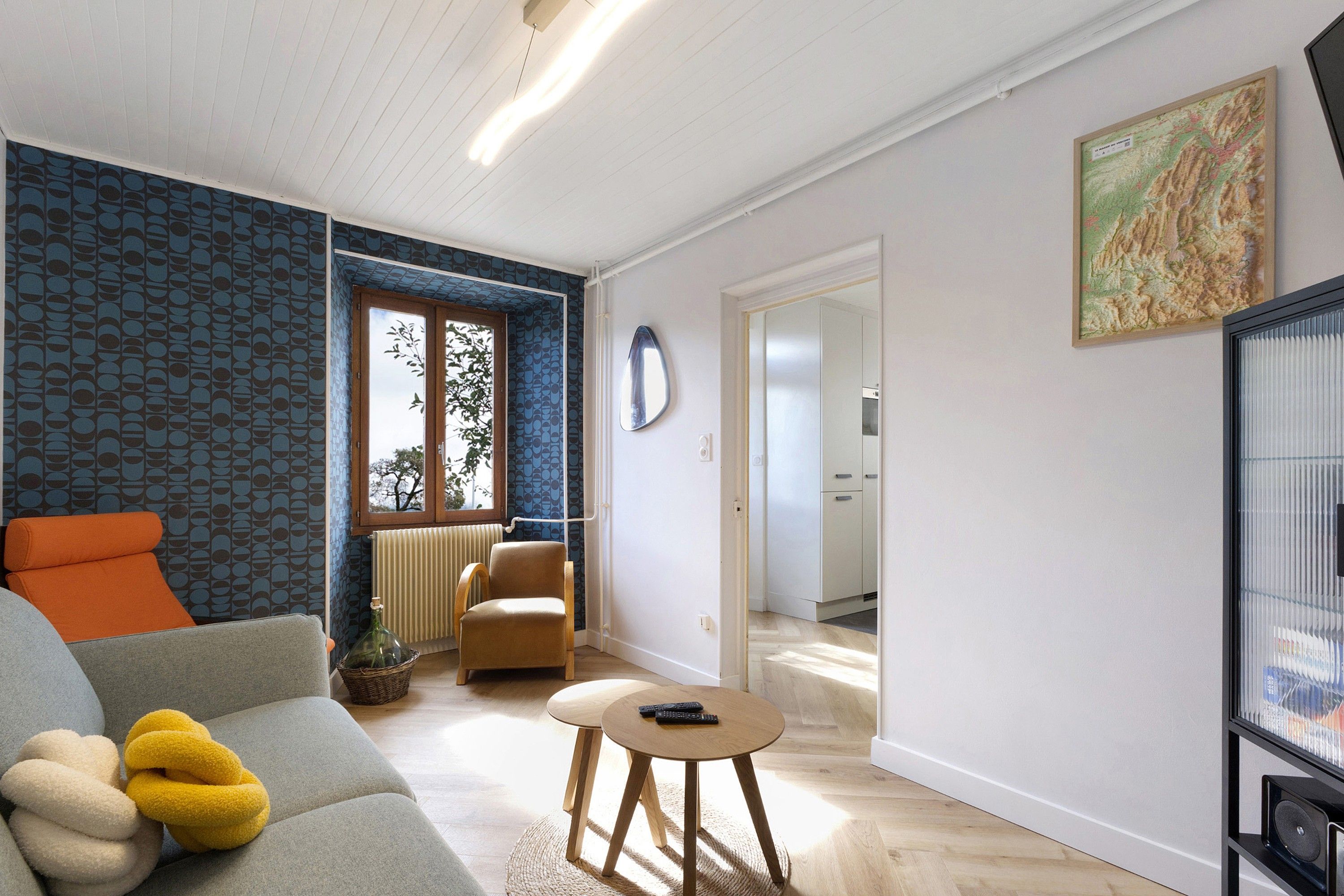 Un salon moderne avec un canapé, un fauteuil et une petite table. Il y a une fenêtre donnant sur des arbres, un radiateur et une porte menant à une autre pièce.
