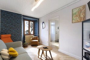 Sala de estar moderna com sofá, poltrona e mesinha. Há uma janela com vista para árvores, um radiador e uma porta que dá para outro cômodo.
