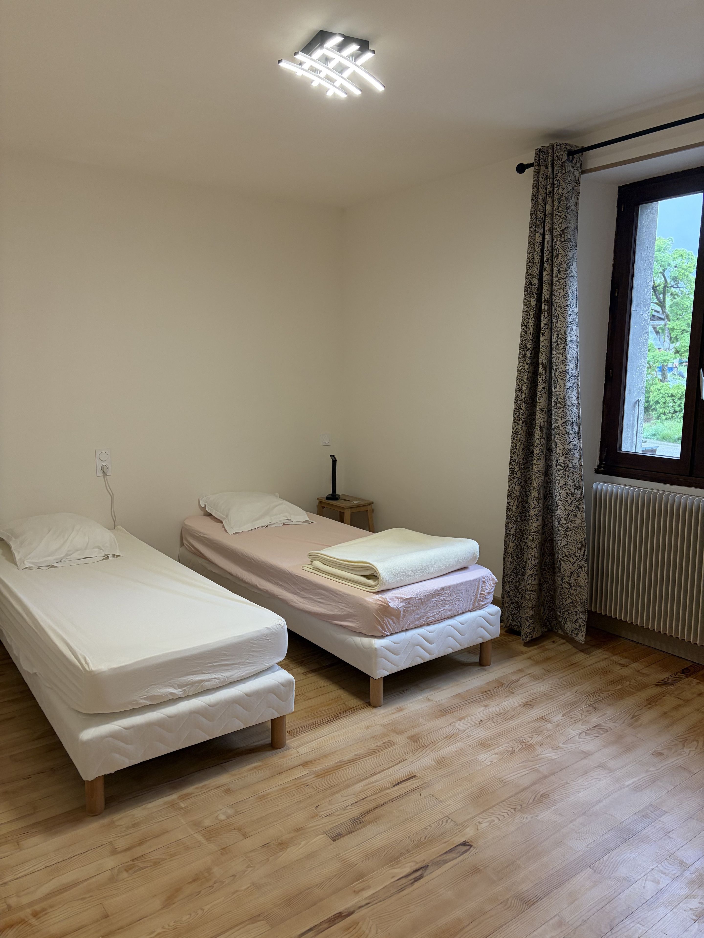 A room with two single beds, a window with curtains, a radiator, and a ceiling light fixture.