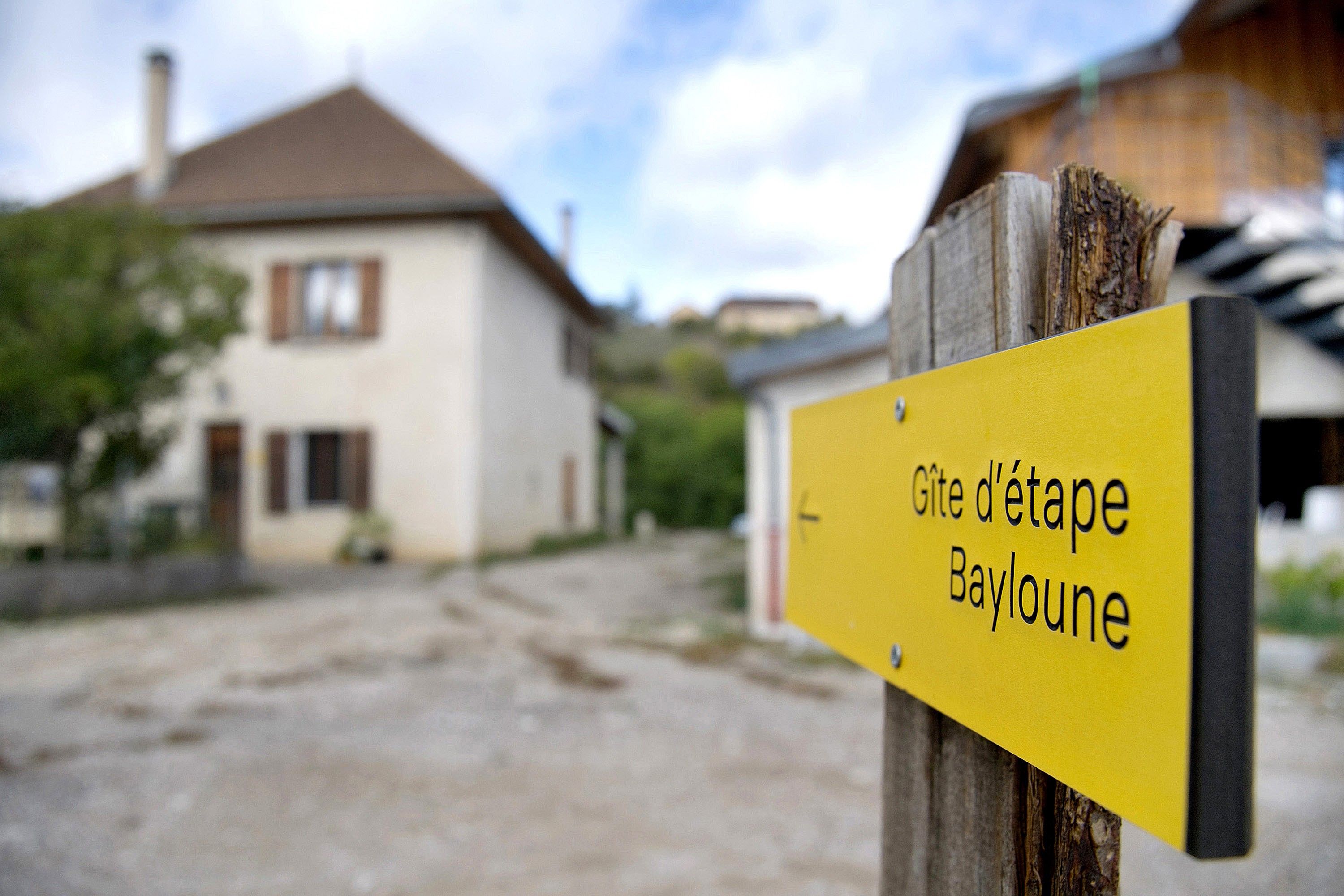 Een geel bord met zwarte letters waarop 'Gîte d'étape Baylounge' staat, voor een gebouw met een bruin dak en witte muren.
