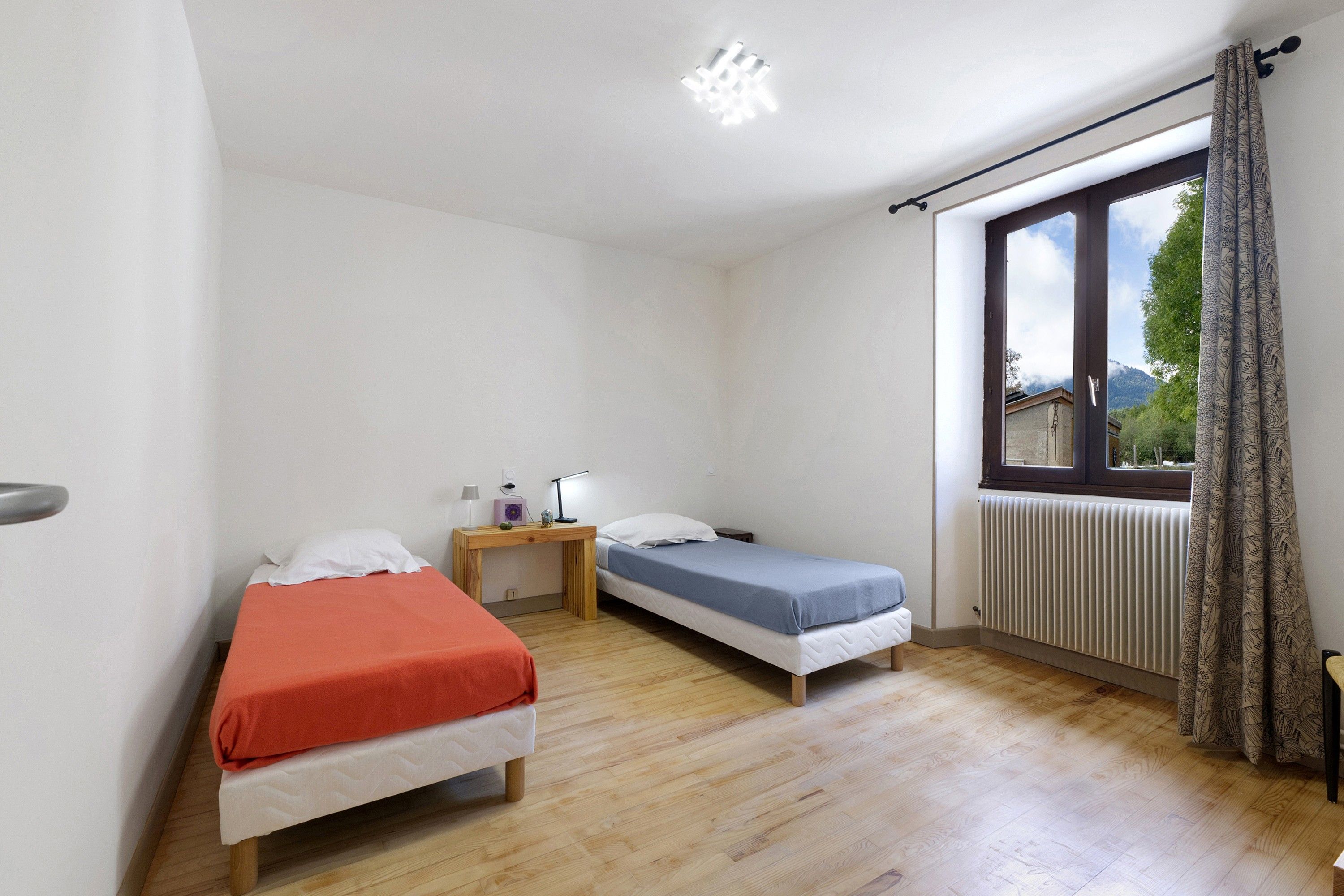 Ein Zimmer mit zwei Einzelbetten, einem kleinen Holztisch mit Lampe, einem Heizkörper und einem Fenster mit Vorhängen.