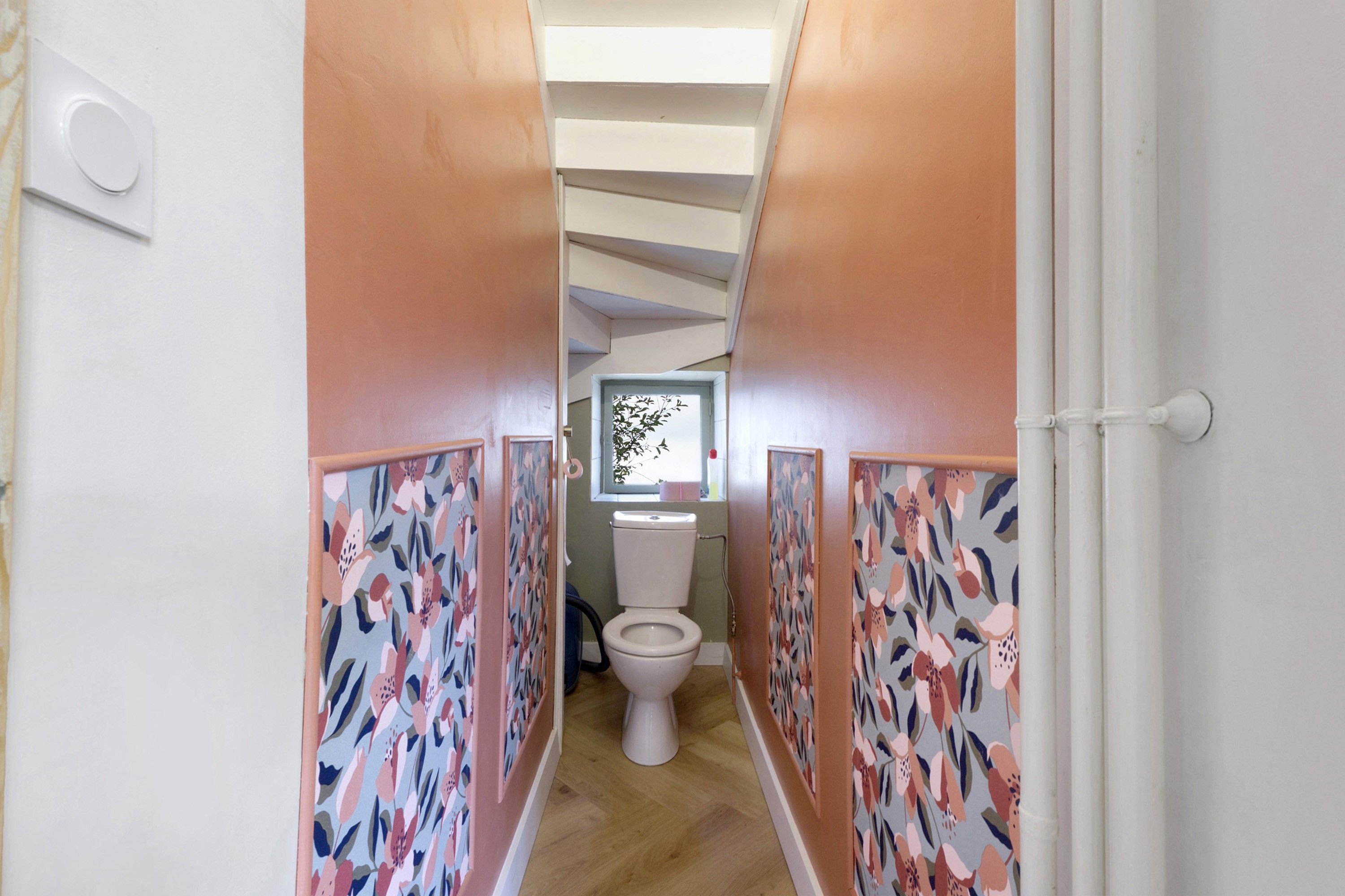 Een smalle gang met behang met patroon, een witte deur aan de rechterkant, en een toilet in de verte.