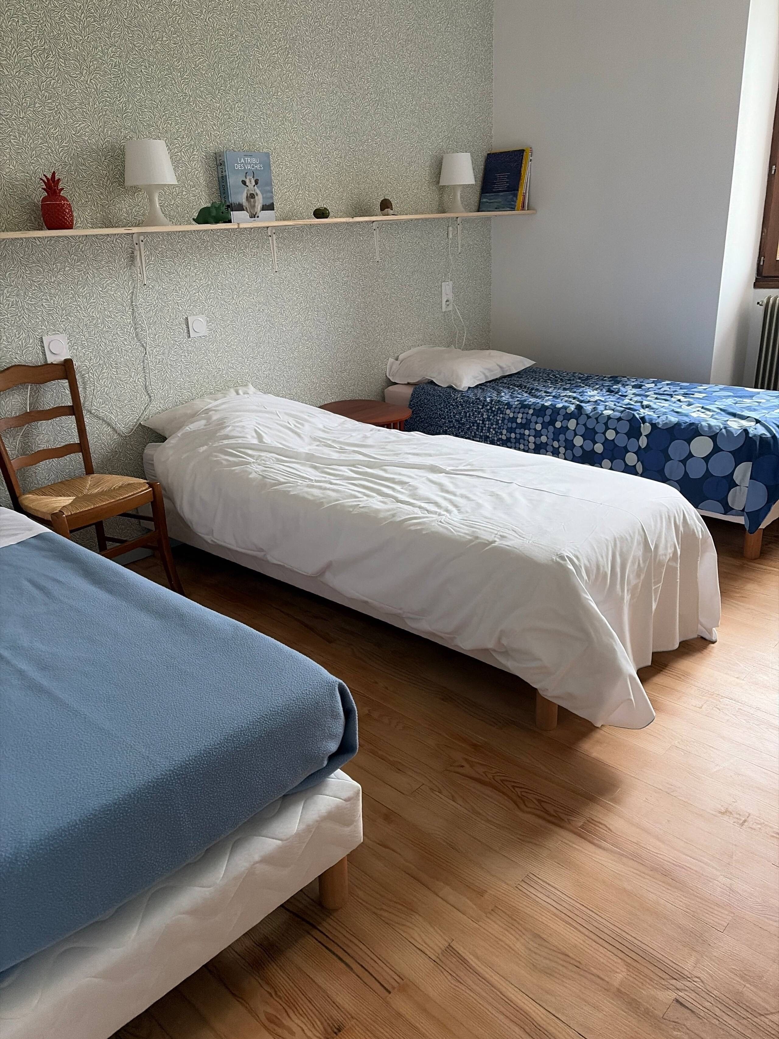 A room with two single beds, a wooden chair, a shelf with books and decorative items, and a patterned wall.