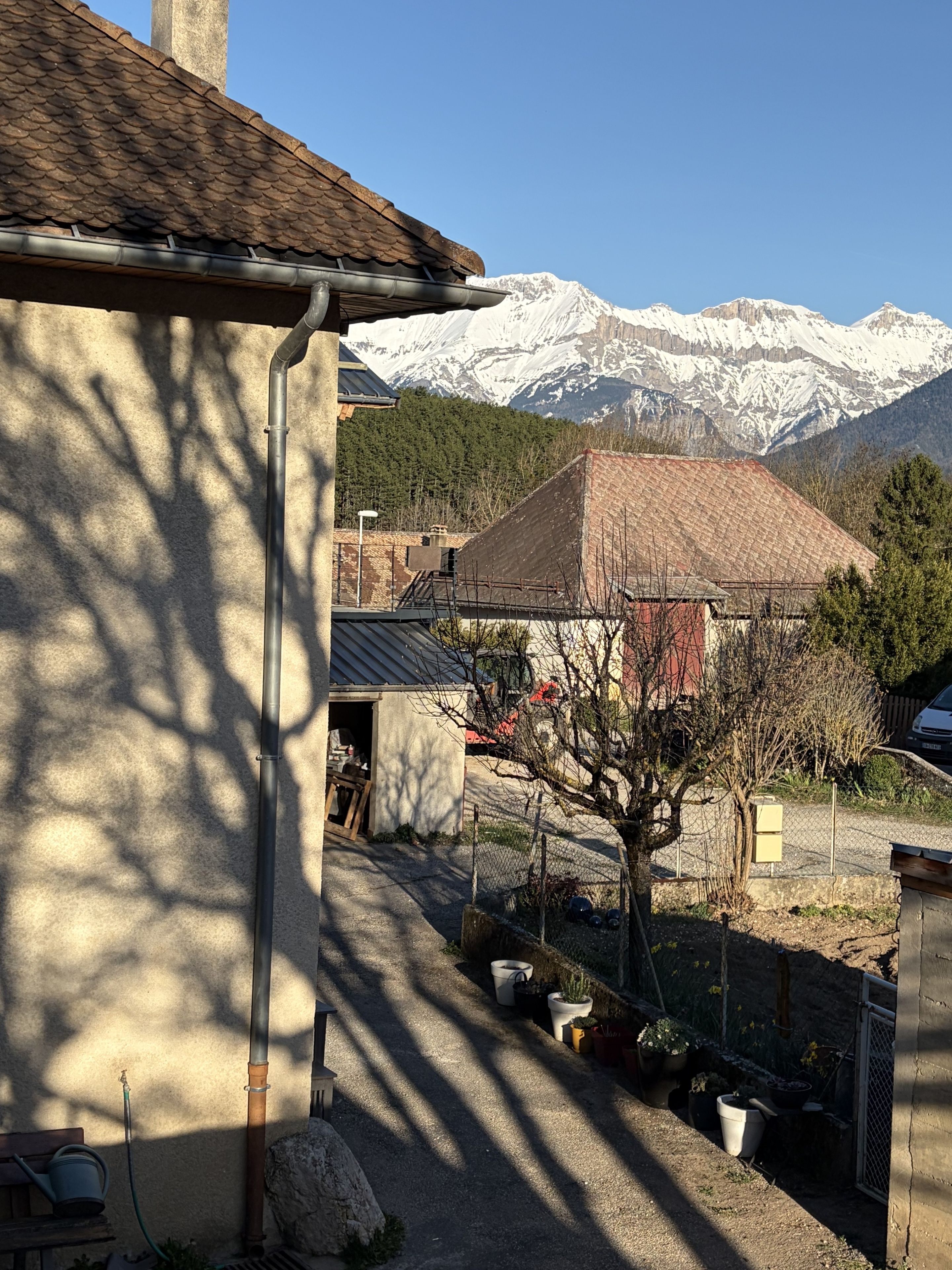 Un bâtiment avec un toit recouvert de tuiles, un jardin agrémenté de plantes en pot et, en arrière-plan, une vue sur des montagnes enneigées.