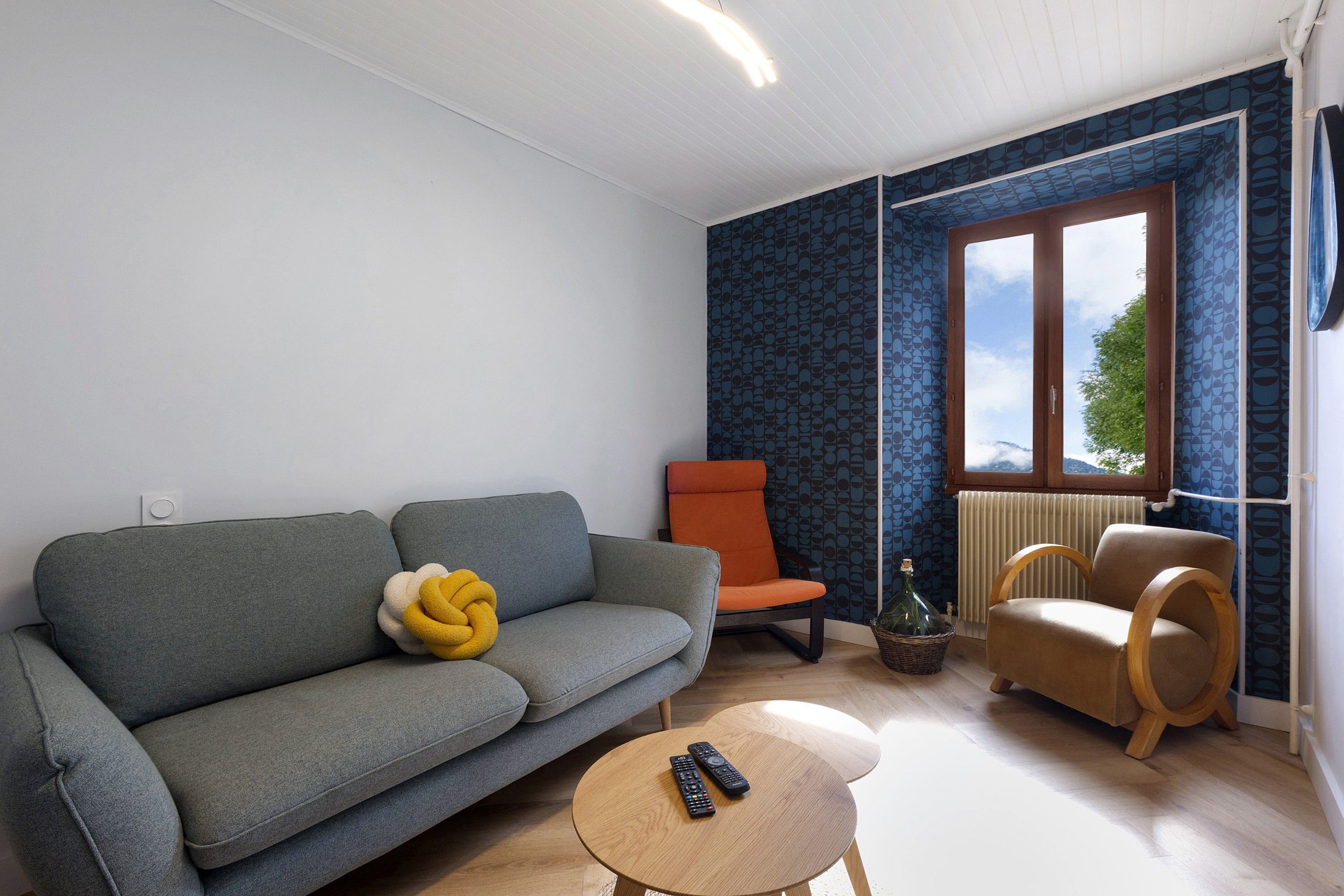 Ein Wohnzimmer mit einem grauen Sofa, einem orangefarbenen Sessel, einem Holzstuhl und einem runden Holztisch mit zwei Fernbedienungen.