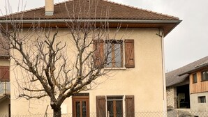 A two-story house with a brown door and shuttered windows, surrounded by a chain-link fence and a tree with bare branches. - Holiday Home 'Étape À Monestier Du Percy' with Mountain View and Private Terrace (Monestier-du-Percy)