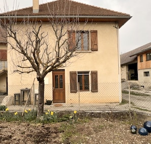 A two-story house with a brown door and shuttered windows, surrounded by a chain-link fence and a tree with bare branches. - Holiday Home 'Étape À Monestier Du Percy' with Mountain View and Private Terrace (Monestier-du-Percy)