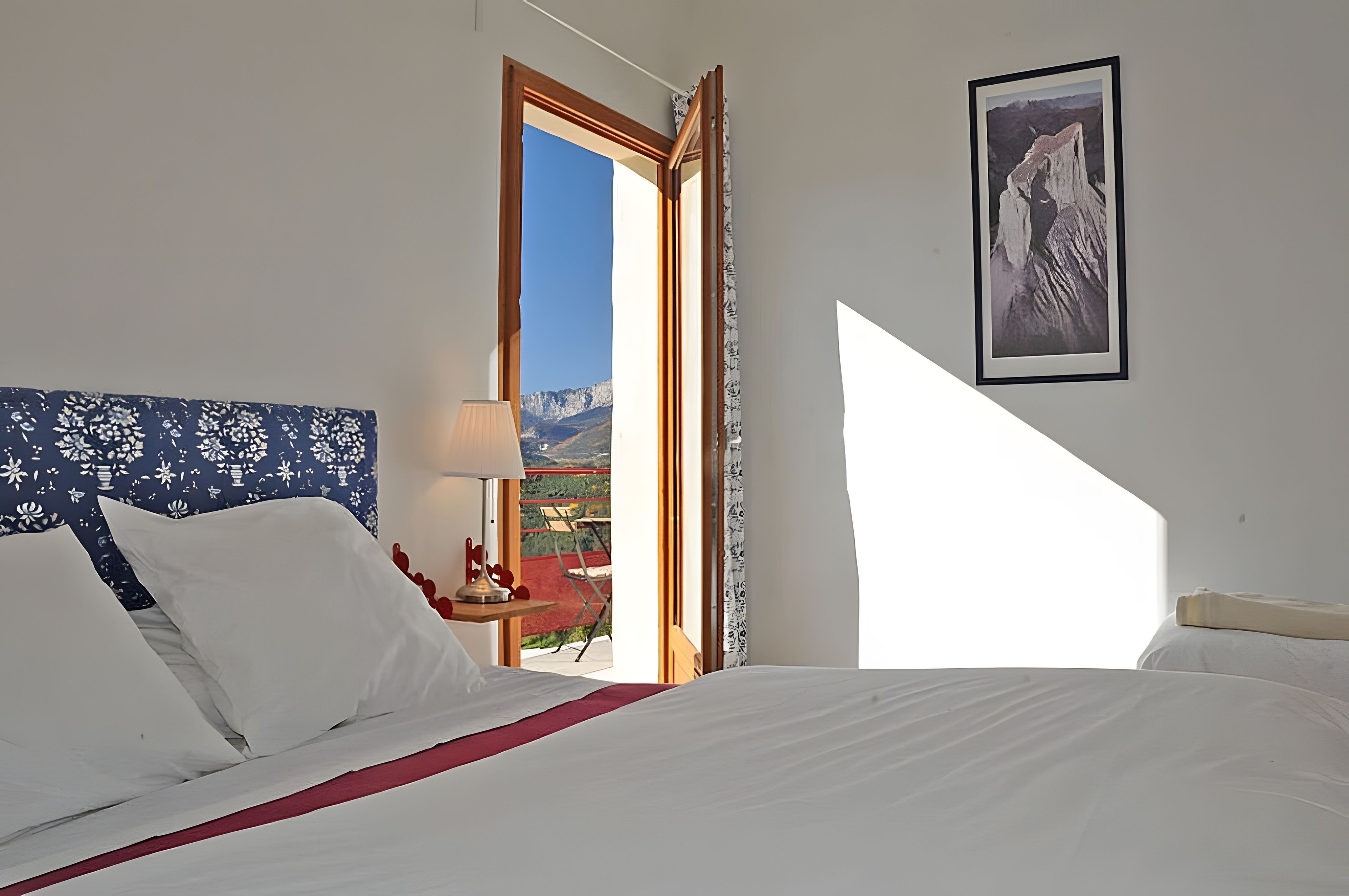 A hotel room with a bed, a window with a mountain view, a framed picture on the wall, and a red chair outside.