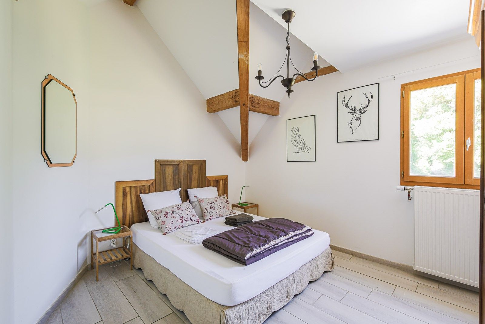 Un dormitorio con techo inclinado, cabecera de madera, cama con colchón blanco y manta morada, mesita de noche con lámpara y cuadros en la pared.