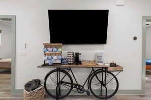 A room with a television mounted on the wall, a coffee station with a coffee maker, a basket, and a tray with mugs.