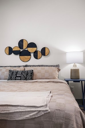 Ein Schlafzimmer mit einem Bett, Kissen, einer Wanddekoration, einer Nachttischlampe und einem kleinen Tisch.