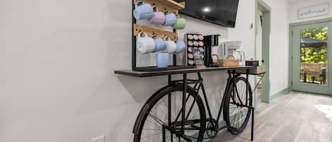 Ein Zimmer mit einem stationären Fahrrad, einer Kaffeestation mit Tassen und einer Kaffeemaschine sowie einem an der Wand montierten Fernseher.