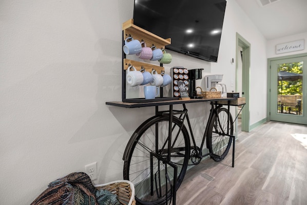 A room with a stationary bike, a coffee station with mugs and a coffee machine, and a television mounted on the wall.