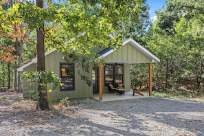A small, single-story cabin with a porch, surrounded by trees.