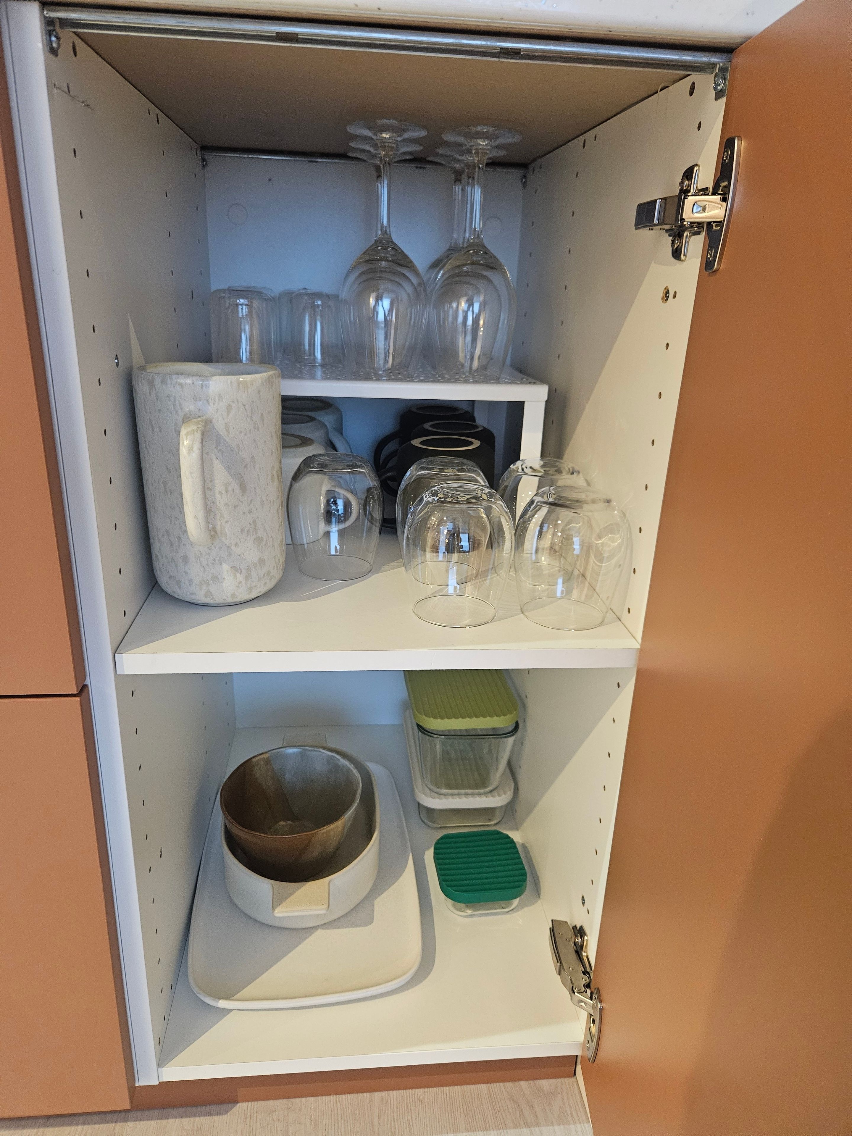 A cabinet with shelves containing glassware, a ceramic mug, a bowl, and a green container.